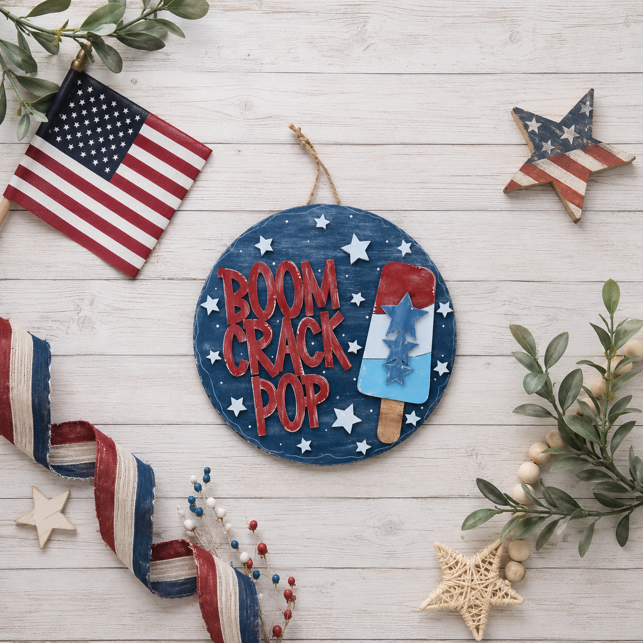 Hand-painted Boom Crack Pop door round with star popsicle design displayed on a wood surface surrounded by an American flag and patriotic ribbon