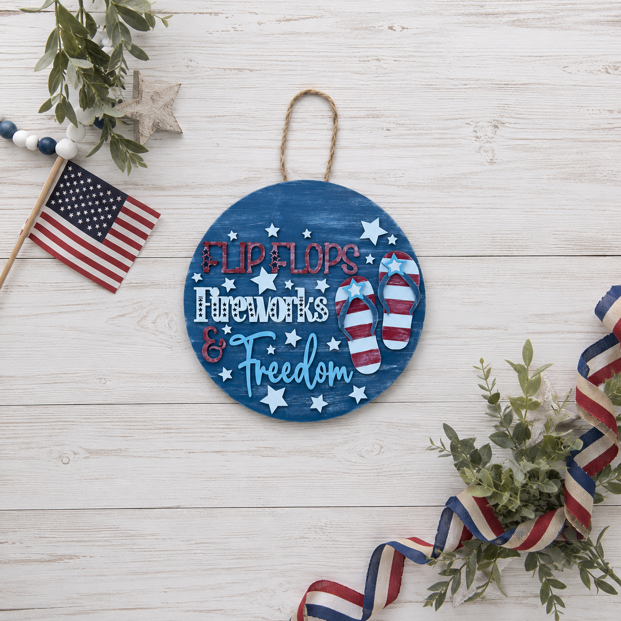 Hand-painted Flip Flops Fireworks and Freedom door round with flip flop design displayed on a wood surface surrounded by an American flag and greenery