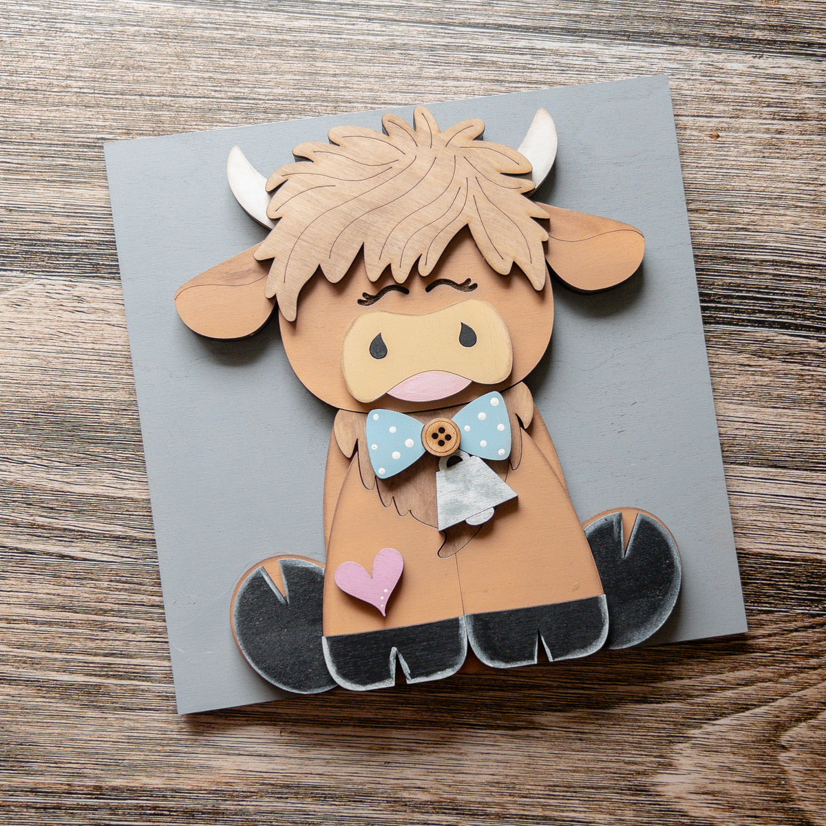 Highland cow porch decor tile with a blue polka-dot bow, silver cowbell, and pink heart on a gray tile, Millie & Co.
