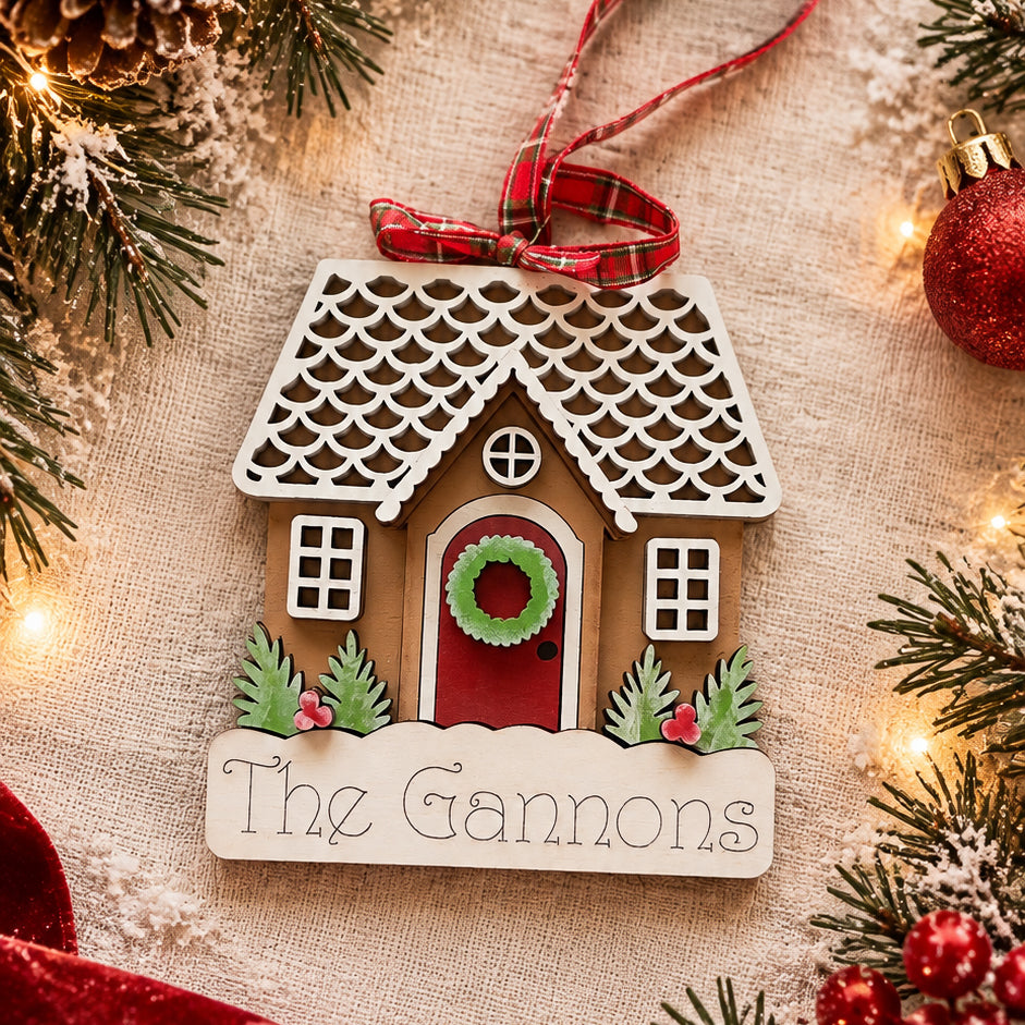 Personalized gingerbread house ornament with family name engraved on snowy base, red door and wreath, hand-painted by Millie & Co.